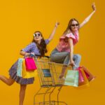 cheerful-happy-girls-with-many-shopping-bags-sitting-shopping-cart-isolated-yellow-studio-background