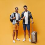 fulllength-portrait-brunet-handsome-men-colorful-shorts-checkered-shirts-white-tshirts-hold-suitcase-tickets-retro-camera-travelers-pose-orange-background