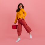 perky-girl-stylish-glasses-stares-amazement-walking-pink-wall-brunette-culottes-orange-blouse-posing-with-red-handbag